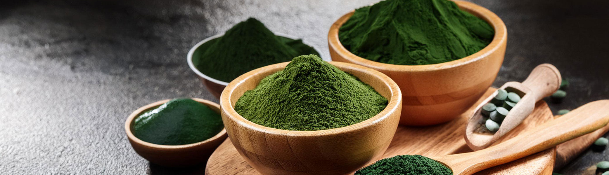 Mit grünem Spirulina-Pulver gefüllte Holzschalen und Löffel sowie Spirulina-Tabletten, die auf einer dunklen Fläche ausgestellt sind.