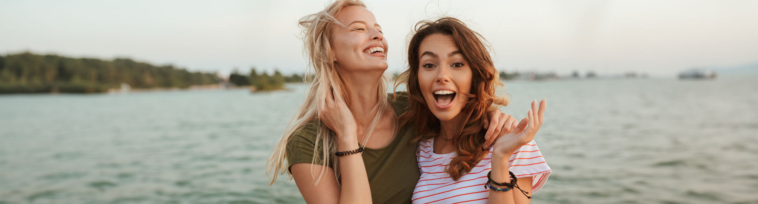 Zwei junge Frauen, die lächelnd und lachend am Wasser sitzen, mit Grünzeug und einem dunstigen Horizont im Hintergrund an einem sonnigen Tag.