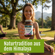 Eine lächelnde Frau sitzt im Gras und hält eine Dose Shilajit von gse.organic.supplements. Im Hintergrund sind Bäume und Berge zu sehen, die an die Reinheit des Himalayas erinnern. Deutscher Text: Naturtradition aus dem Himalaya.