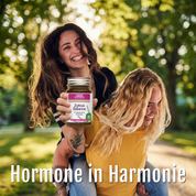 Zwei lächelnde Frauen im Freien, von denen die eine die andere Huckepack nimmt. Die vordere Frau hält ein Glas mit Zyklus Balance (Bio) von gse Bio-Nahrungsergänzungsmitteln, die das hormonelle Gleichgewicht von Frauen hervorheben. Das Sonnenlicht fällt durch die Bäume. Text: "Hormone in Harmonie".